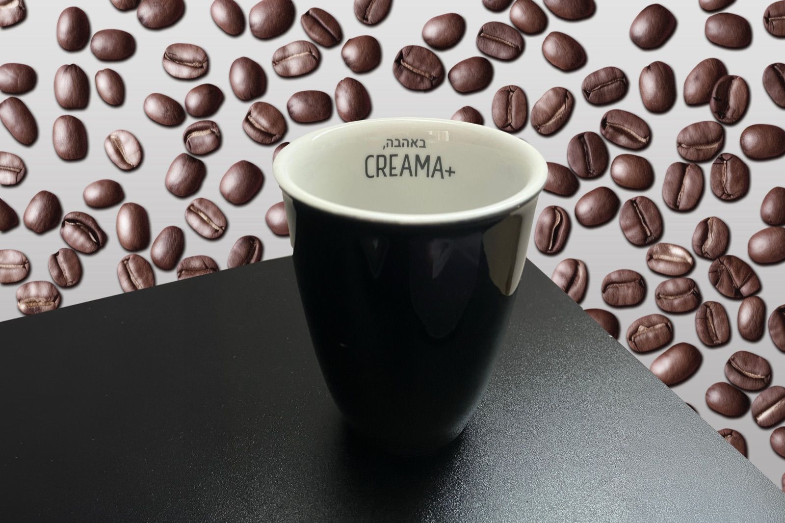 A black branded coffee cup on a dark counter, against a wall of roasted coffee beans